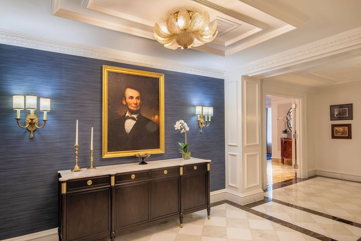 A hallway with a dark blue wall, a marble console table, a framed portrait, wall sconces, and a chandelier.
