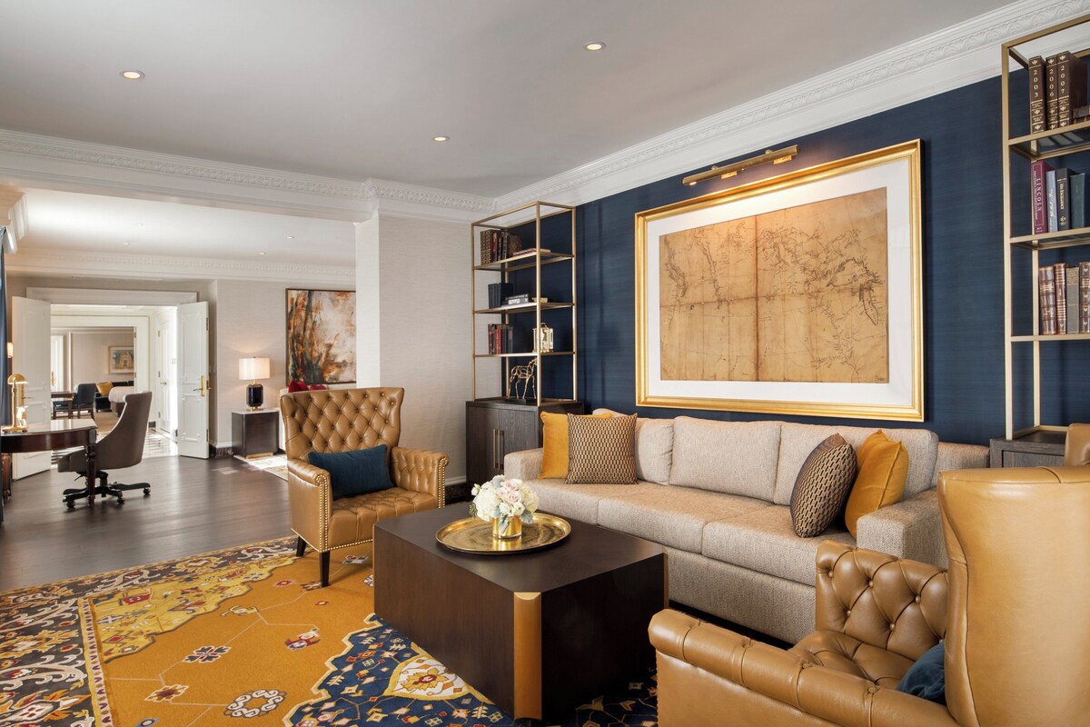 A spacious living room with a large sofa, a coffee table, and a bookshelf. There is a large painting on the wall and a rug on the floor.