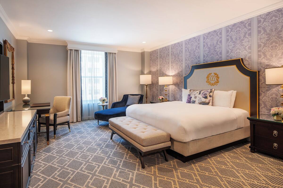 A hotel room with a large bed, a desk, a chair, and a bench.