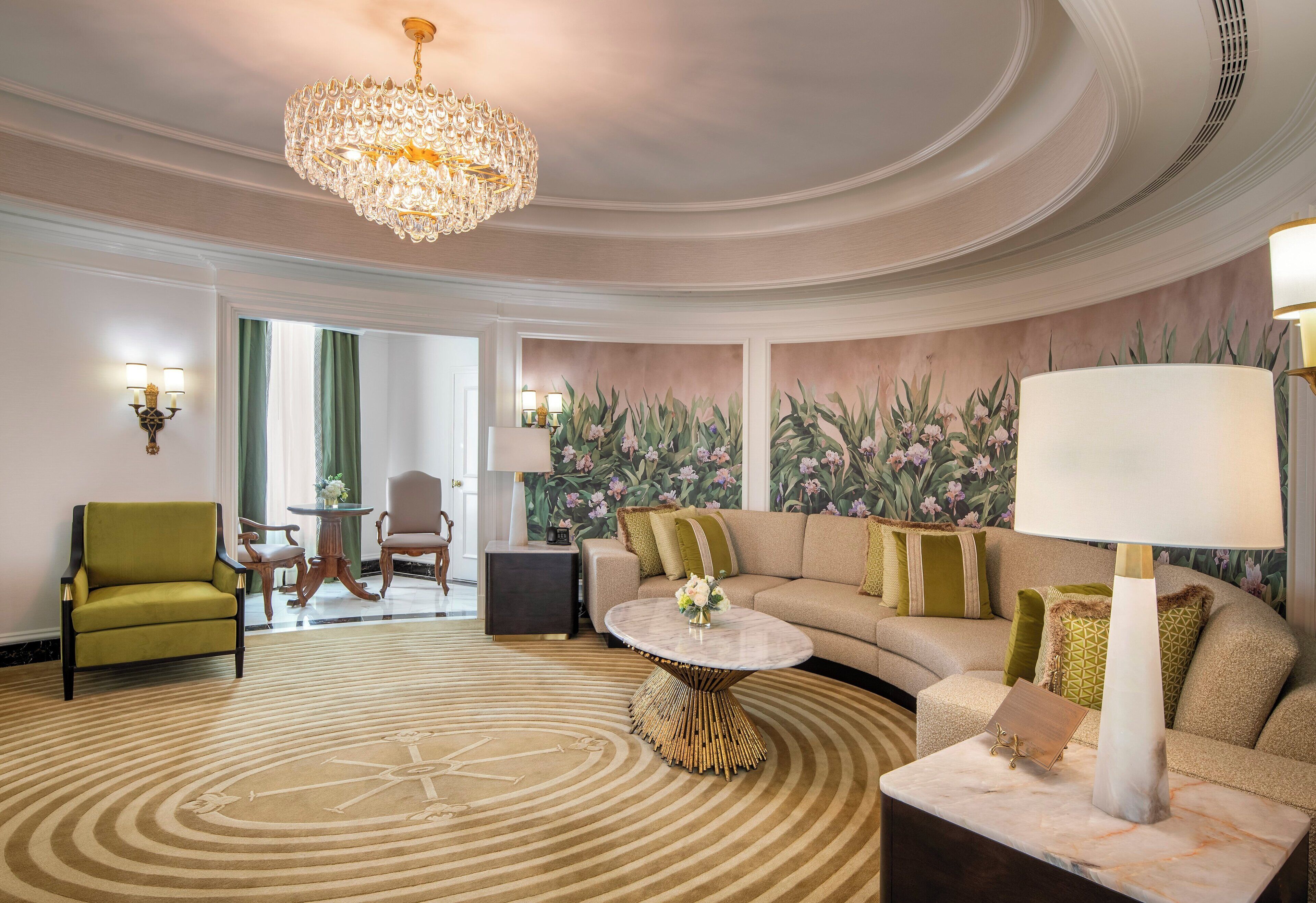 A spacious living room with a large sofa, a round coffee table, and a chandelier. There is a green armchair and a small table with a lamp. The room has a floral wallpaper and a large window with green curtains.