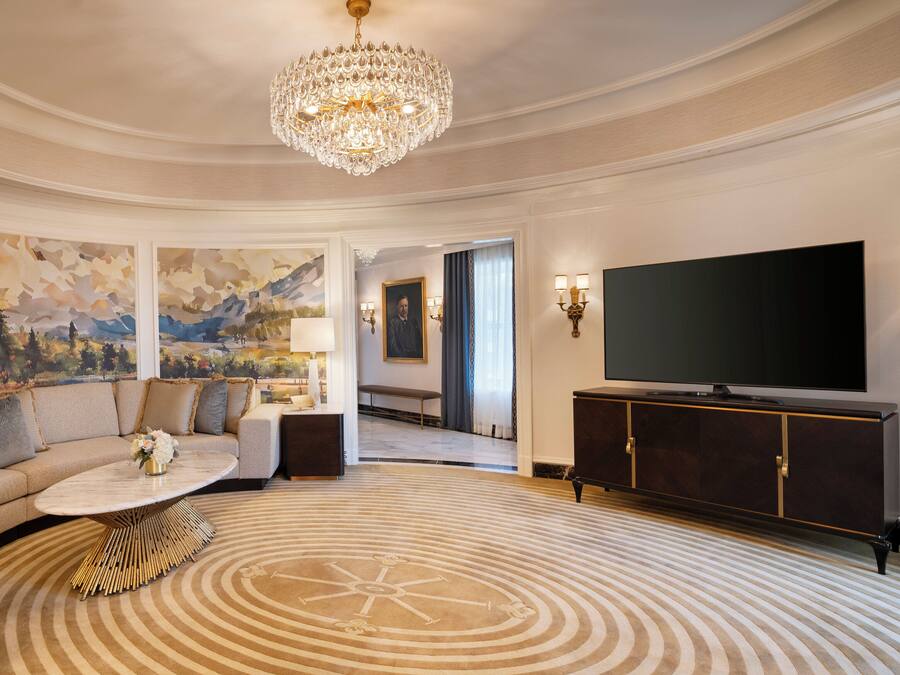 A luxurious living room with a large chandelier, a flat-screen TV, a sofa with decorative pillows, a coffee table, and a patterned carpet.