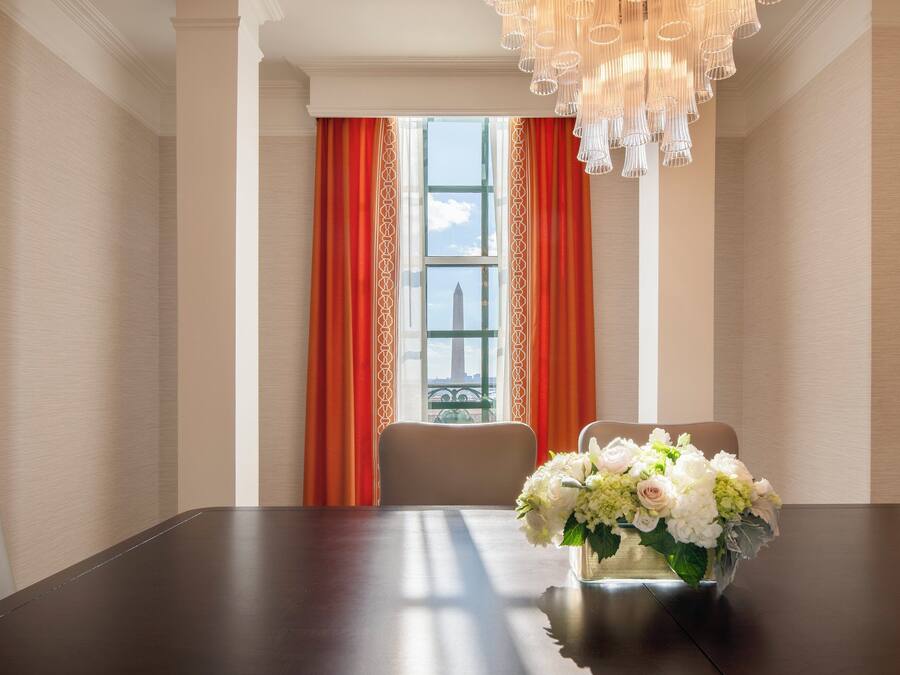 Un salón de conferencias con una mesa de madera oscura, dos sillas, un jarrón de flores y vistas a un edificio a través de una ventana con cortinas rojas.