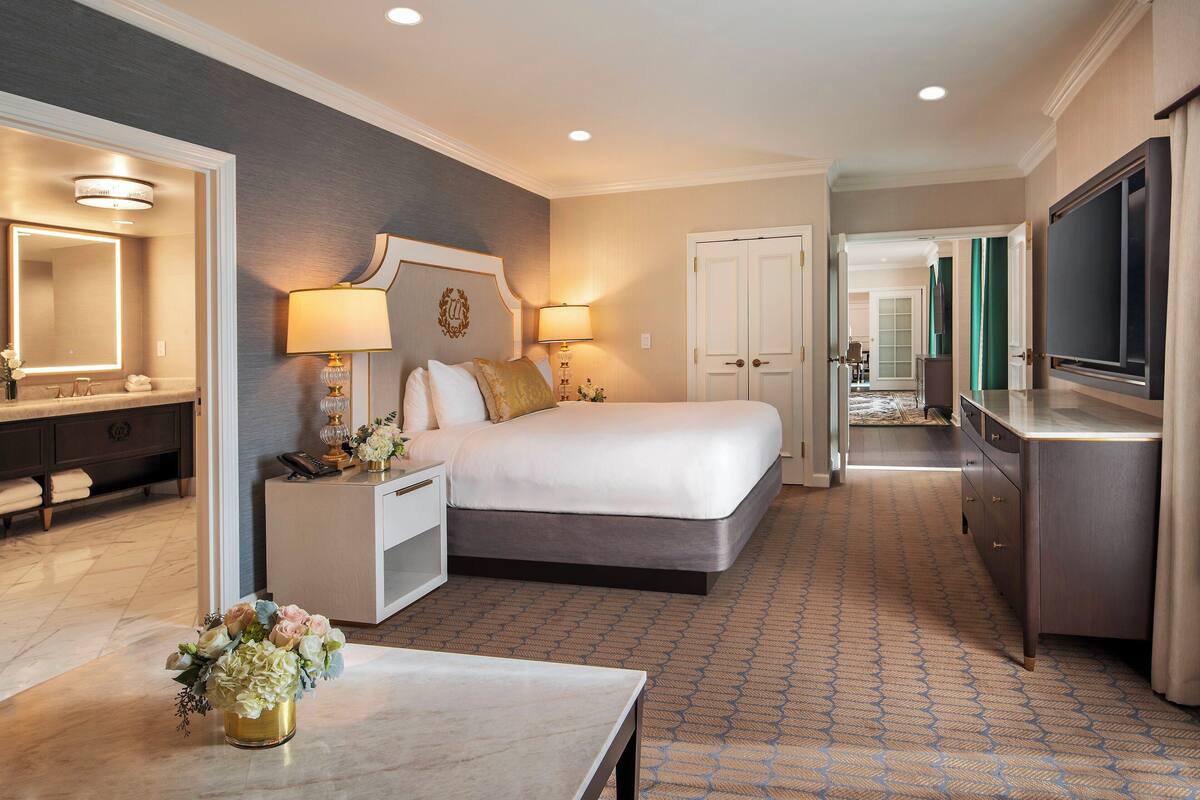A hotel room with a large bed, bedside tables, a desk, a TV, and a bathroom with a mirror and sink.