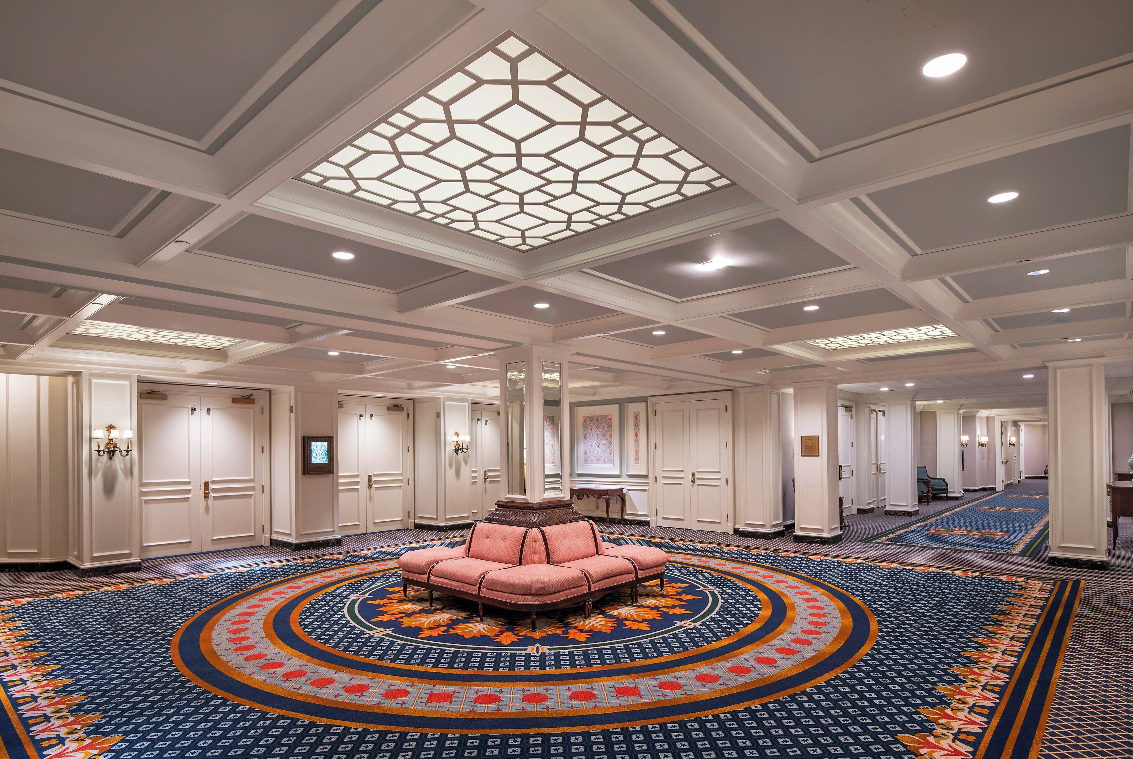 A hotel lobby with a central seating area, a patterned carpet, and a ceiling with a geometric light fixture.