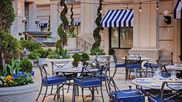 An outdoor dining area with tables and chairs, surrounded by potted plants and a building with striped awnings. - Willard InterContinental Washington by IHG (Washington)