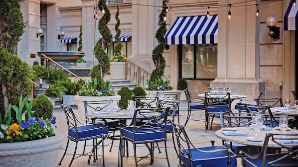 Un espace de restauration en plein air, avec des tables et des chaises, entouré de plantes en pot et bordé d’un bâtiment doté d’auvents à rayures.