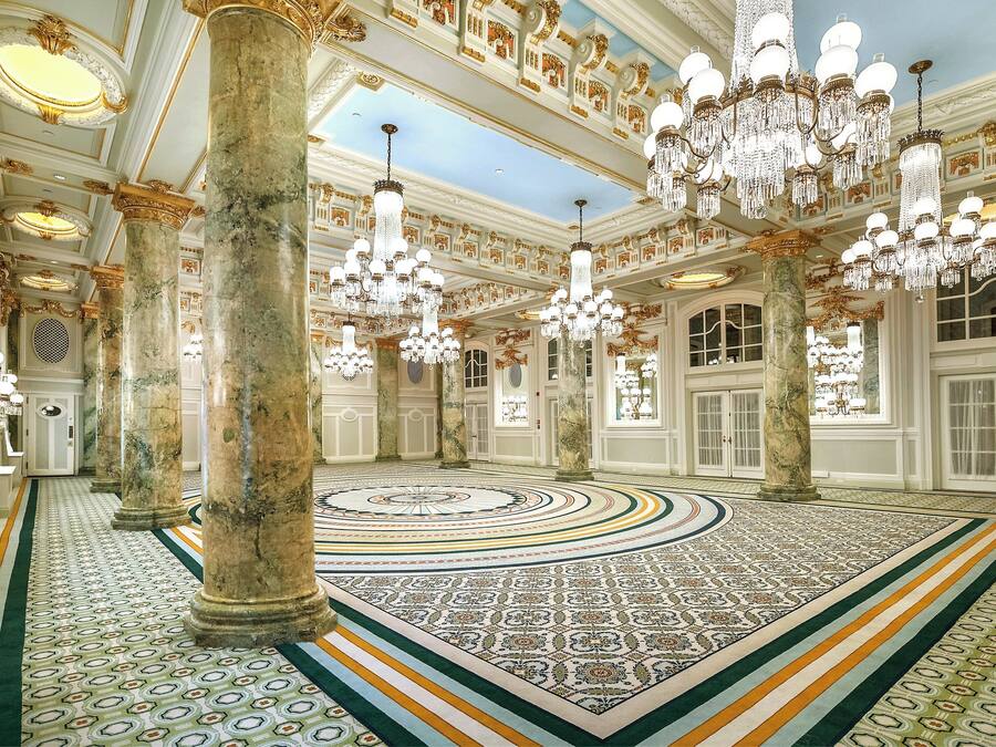 Un gran salón con candelabros ornamentados, columnas de mármol y un suelo con diseño.