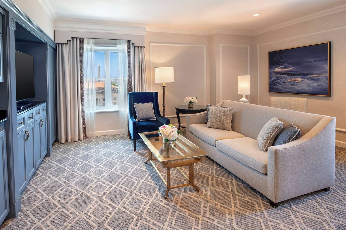 A hotel room with a sofa, a coffee table with flowers, a TV, a lamp, and a painting on the wall.