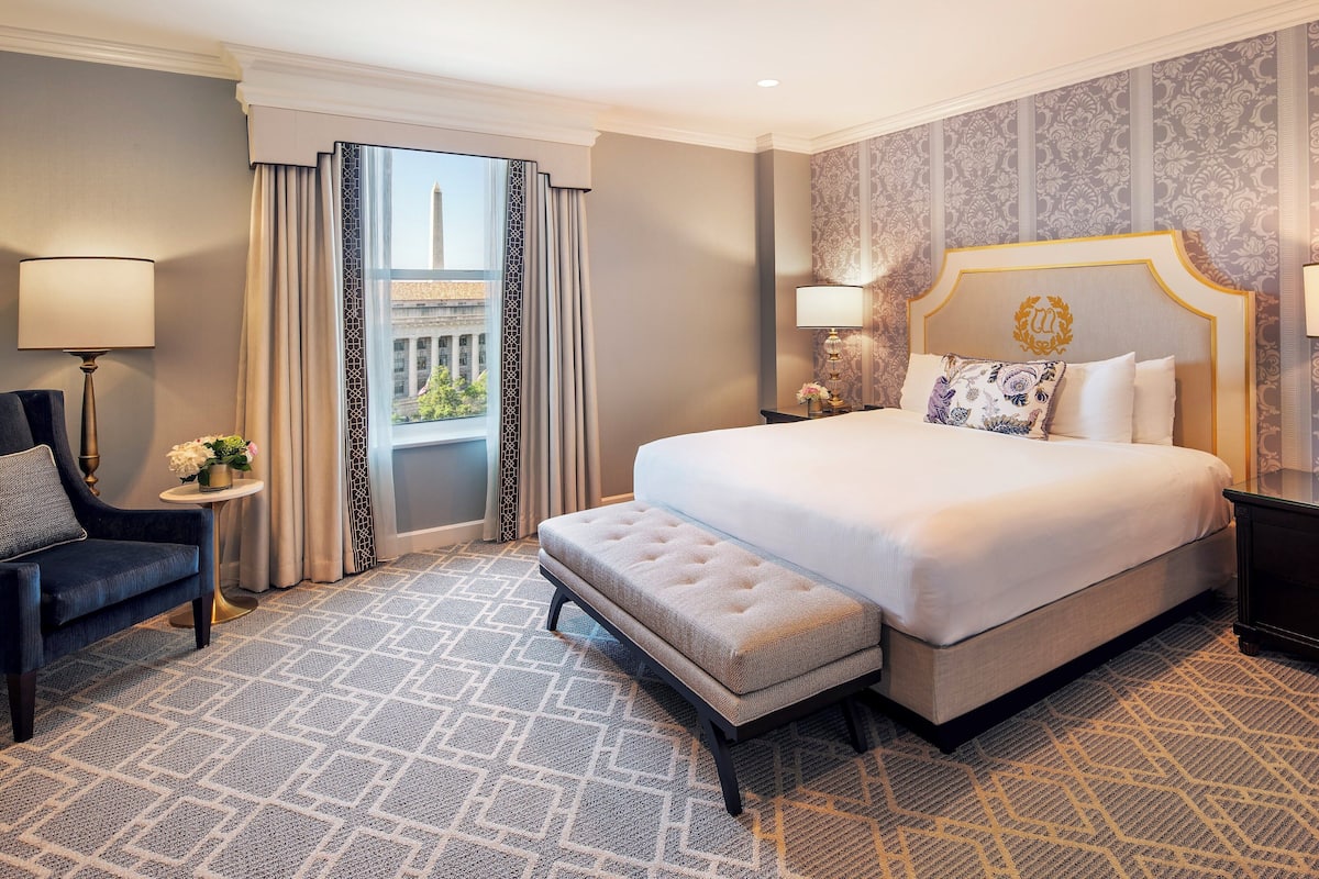 A hotel room with a large bed, a bench, a chair, a nightstand, and a view of a building through a window.