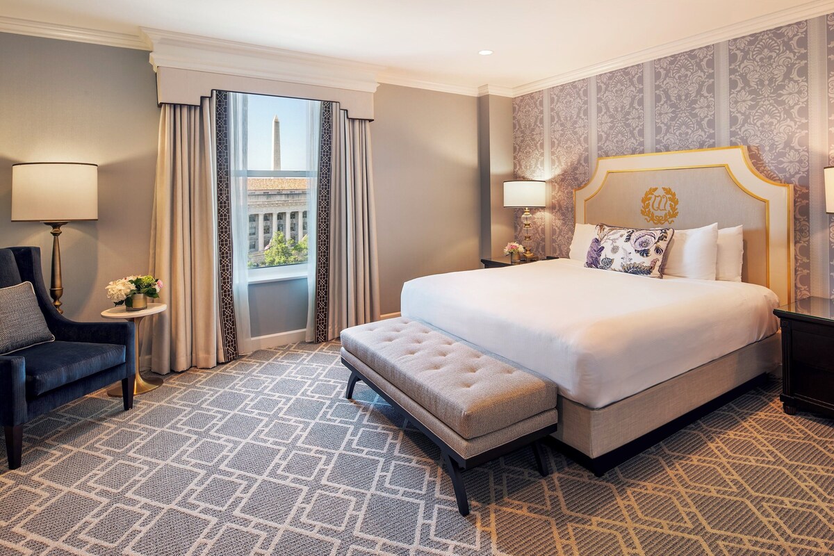 A hotel room with a large bed, a bench, a chair, a nightstand, and a view of a building through a window.