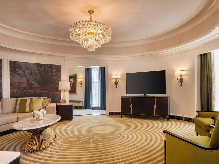 A spacious living room with a large chandelier, a flat-screen TV, and a patterned carpet.