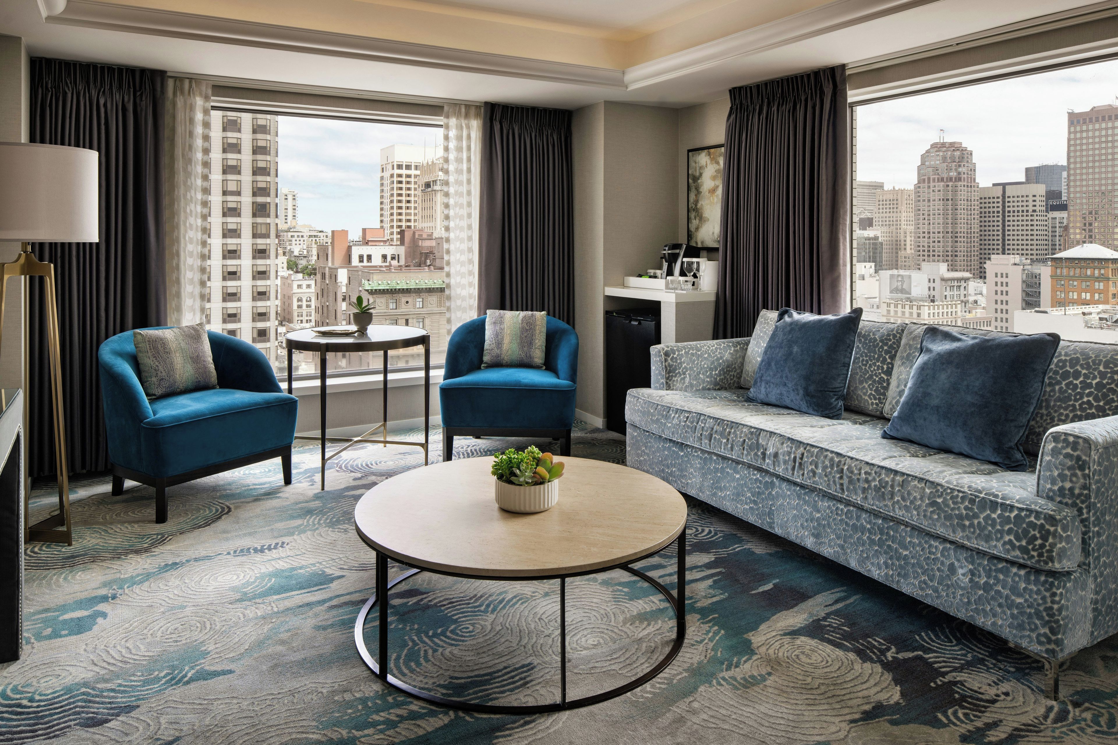 Quarto de hotel moderno com vista para a cidade, um sofá, duas poltronas, uma mesa de centro e uma pequena mesa lateral.