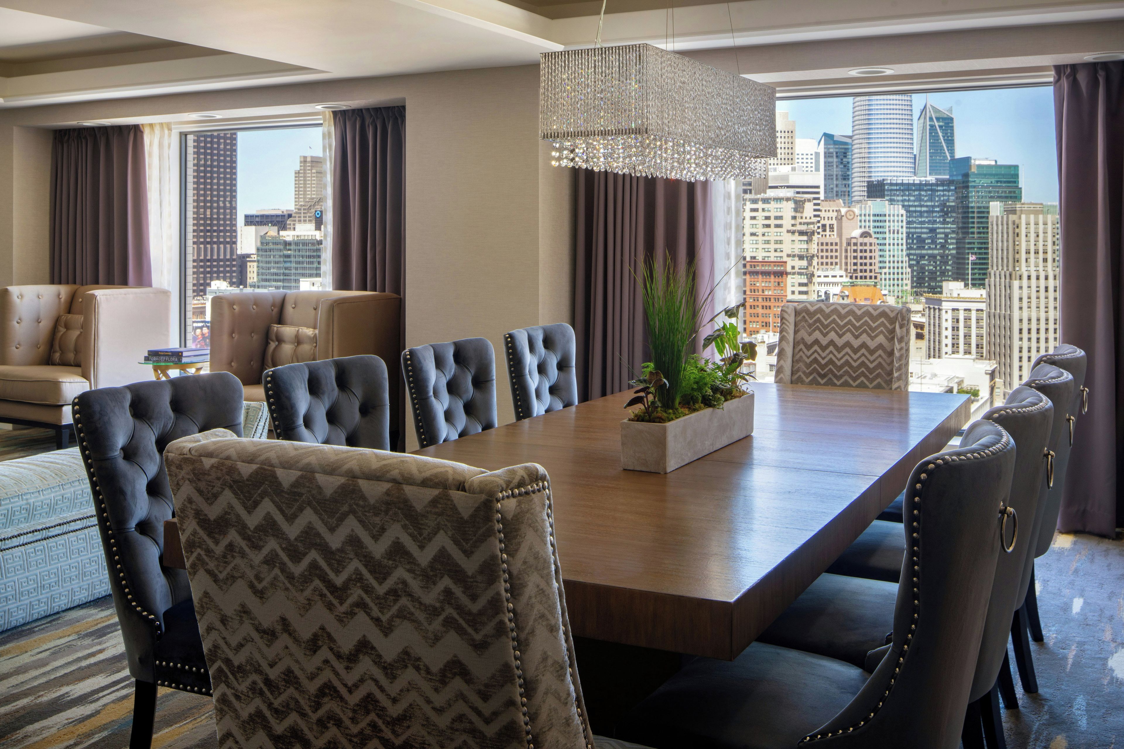 A hotel dining room with a long table, upholstered chairs, a chandelier, and a city view.