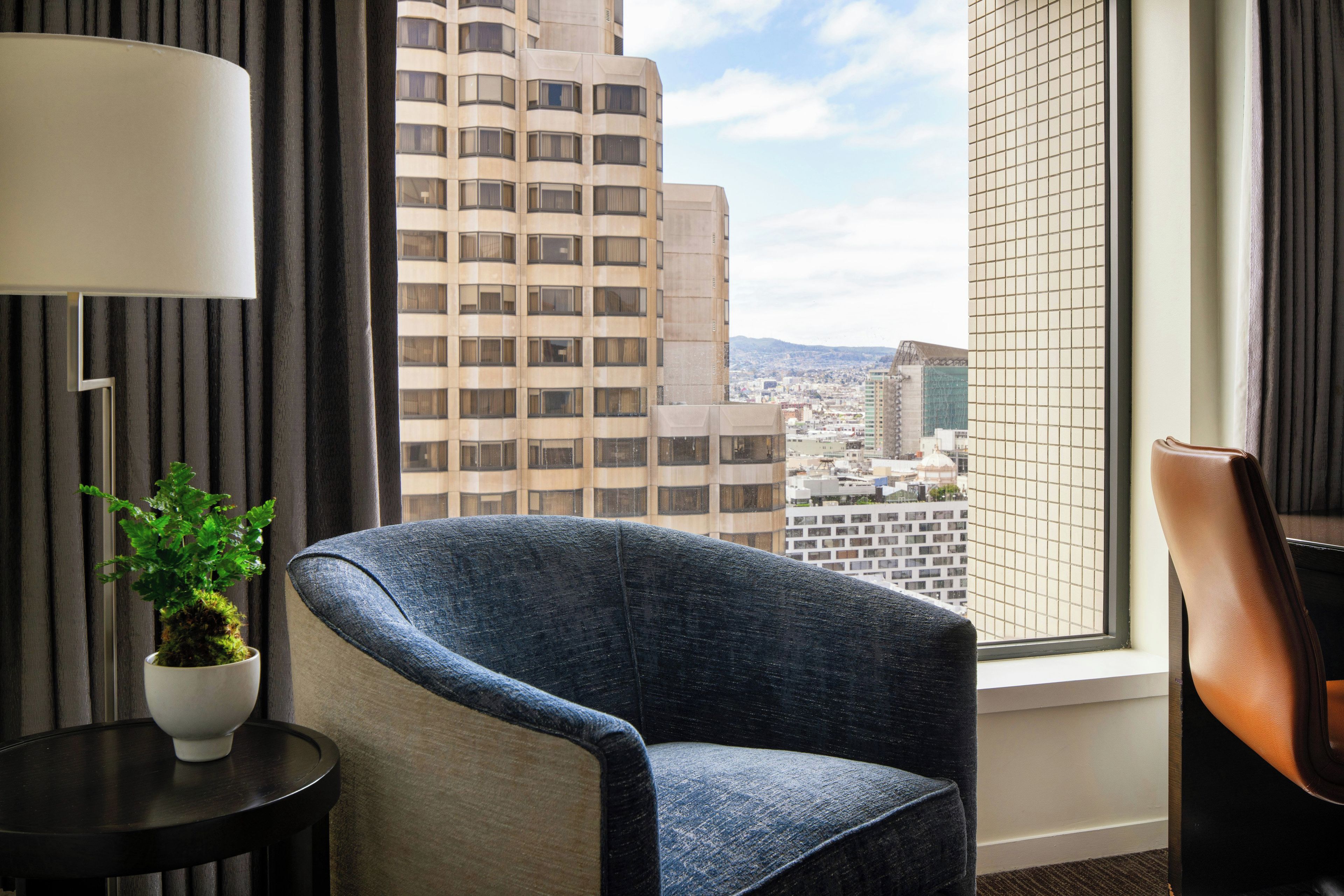 A hotel room with a view of city buildings, a chair, a table with a plant, and a lamp.