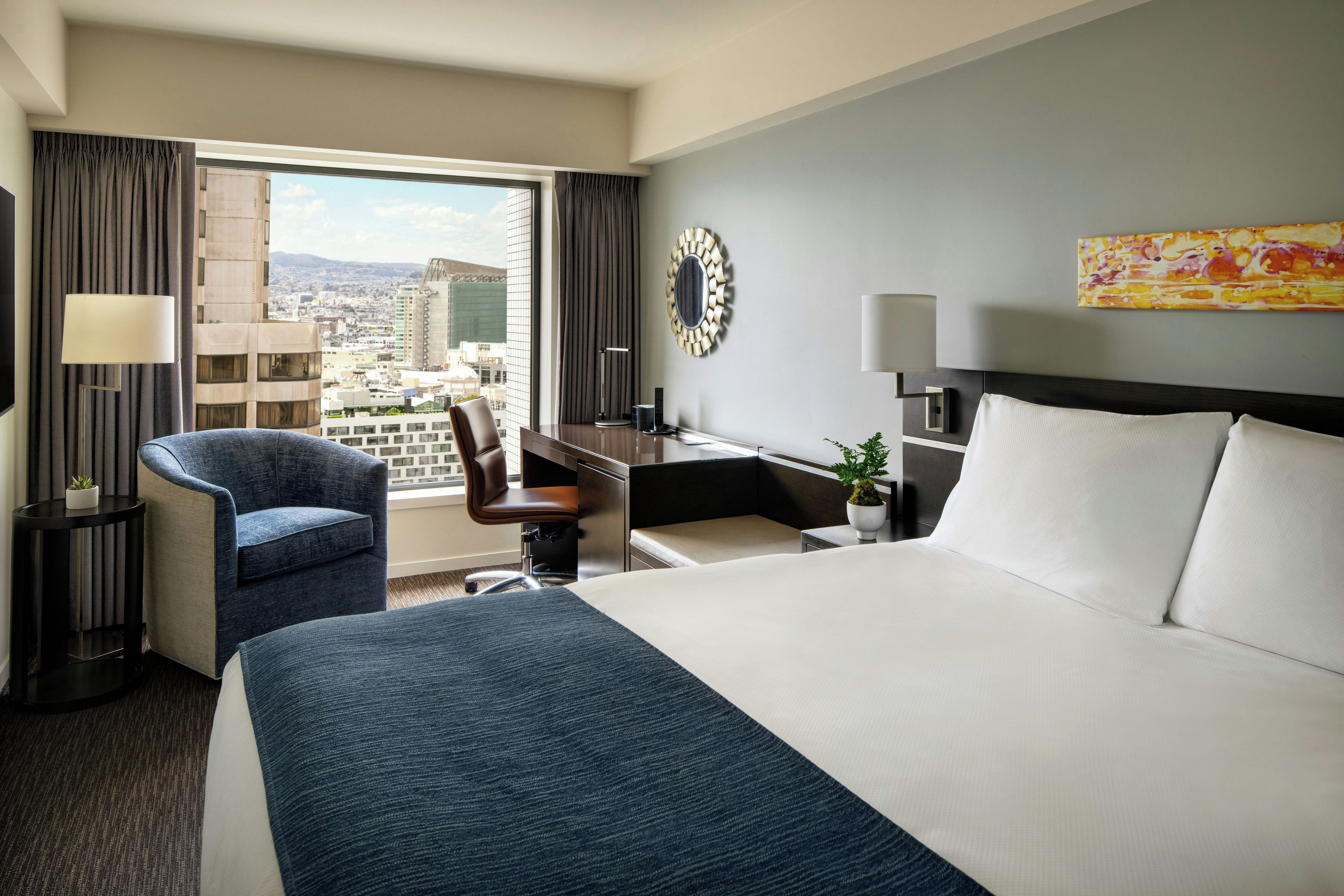 Ein Hotelzimmer mit Bett, Schreibtisch, Stuhl und Blick auf die Stadt, inklusive Vorhängen am Fenster.