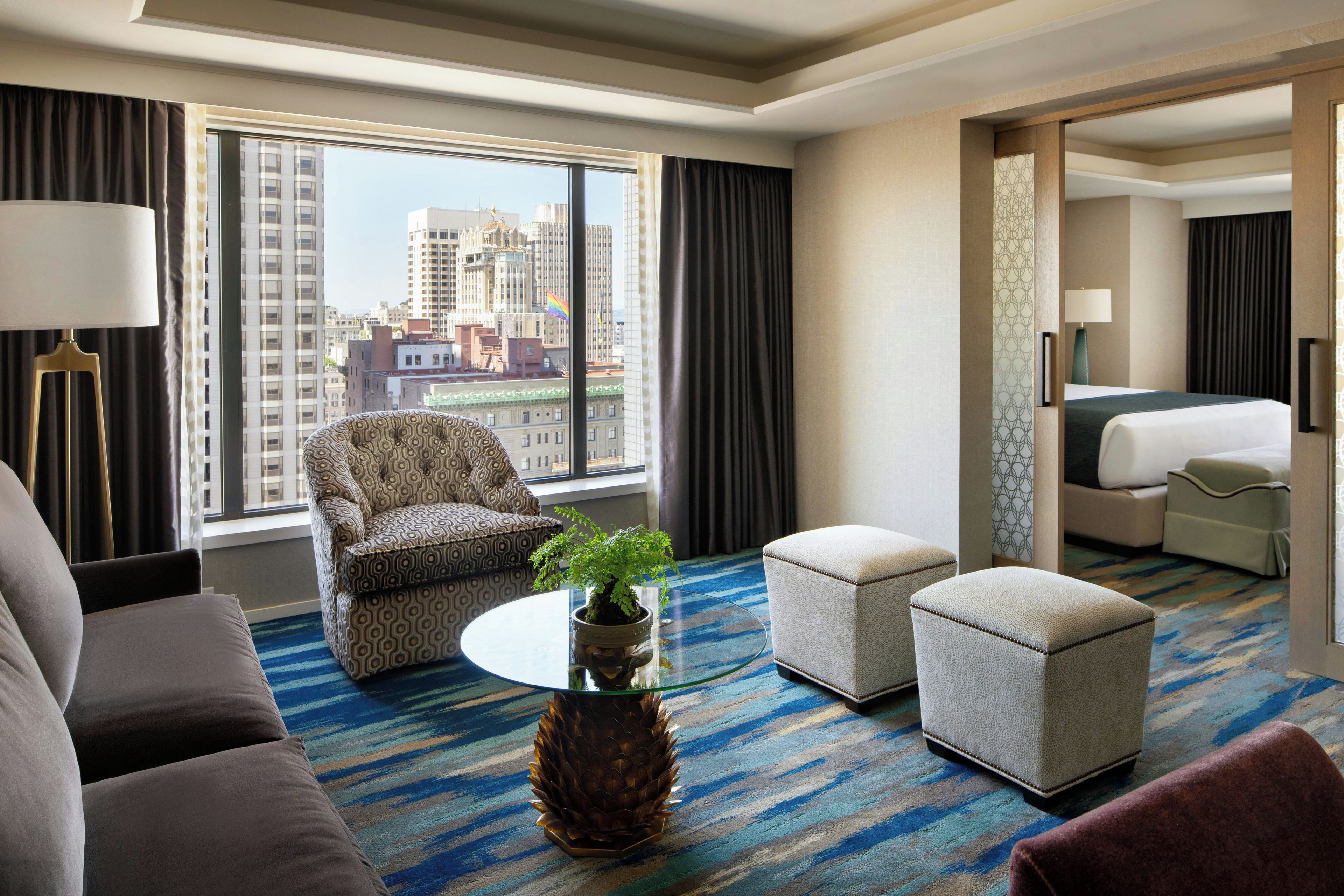 Ein Hotelzimmer mit großem Fenster, einem gemusterten Sessel, einem kleinen Tisch mit Pflanze, zwei Hocker, einer Couch und einem Bett.