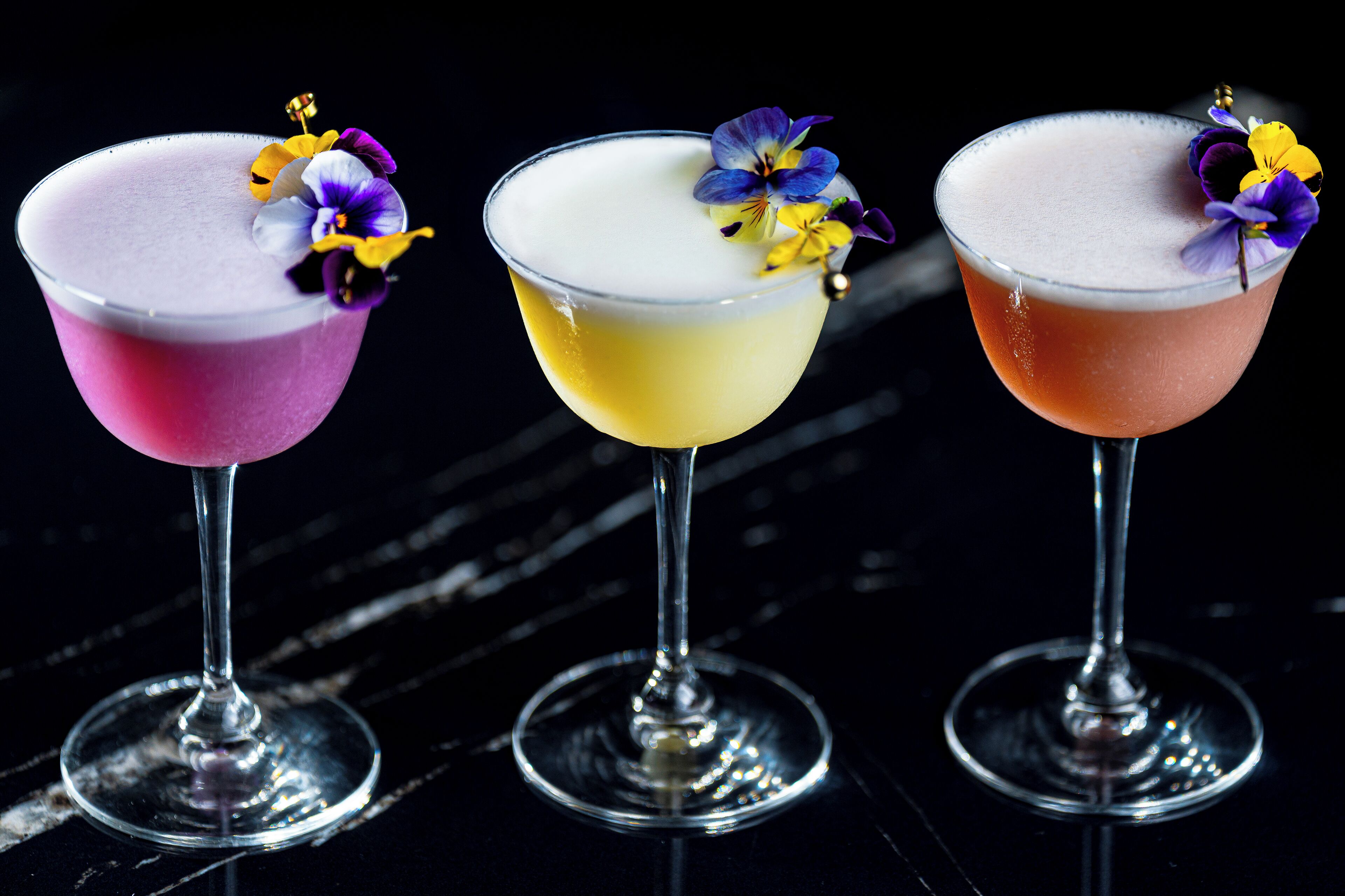 Three cocktails in martini glasses with flowers, on a dark surface.