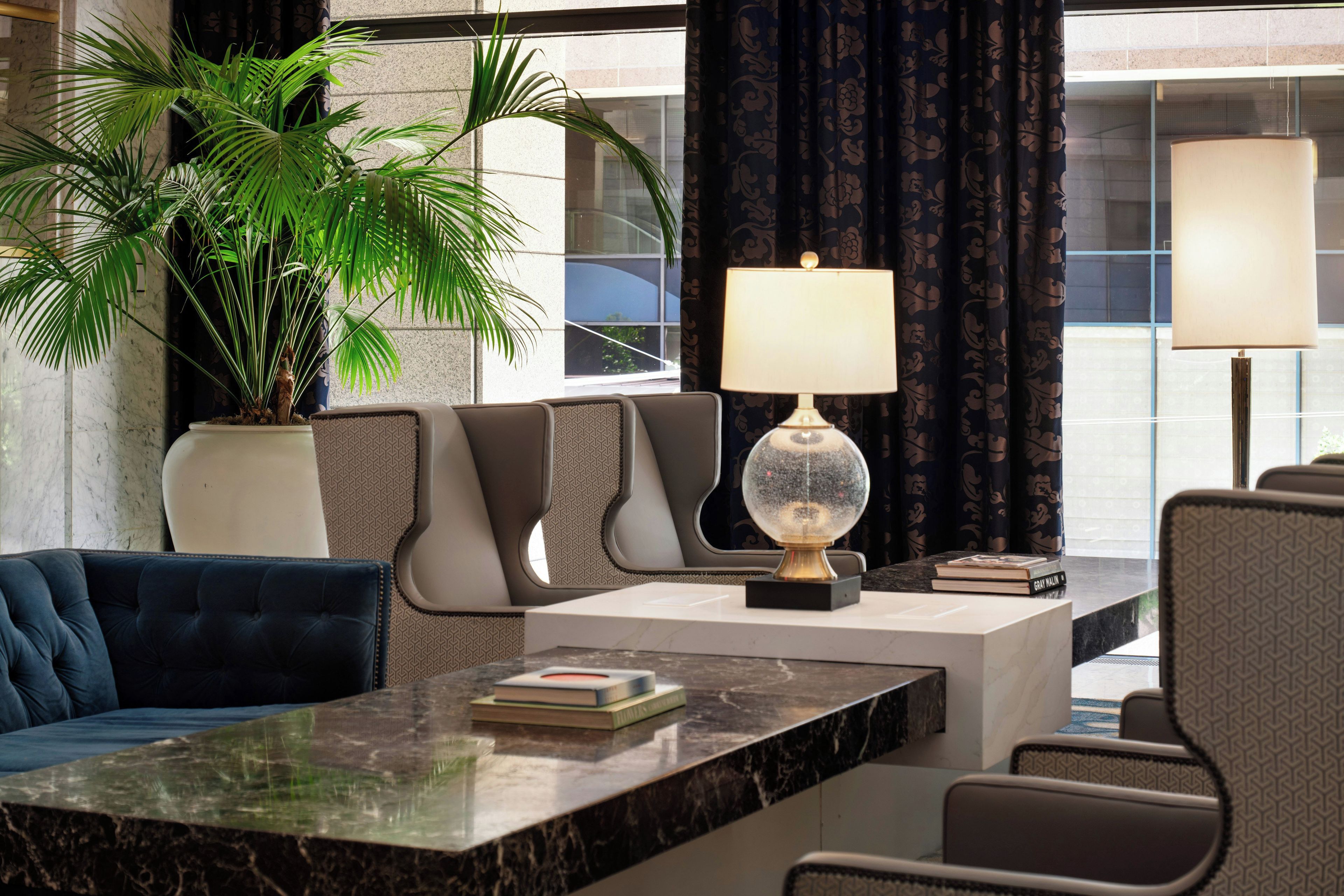 A modern lounge area with a marble table, comfortable seating, a potted plant, and a lamp.