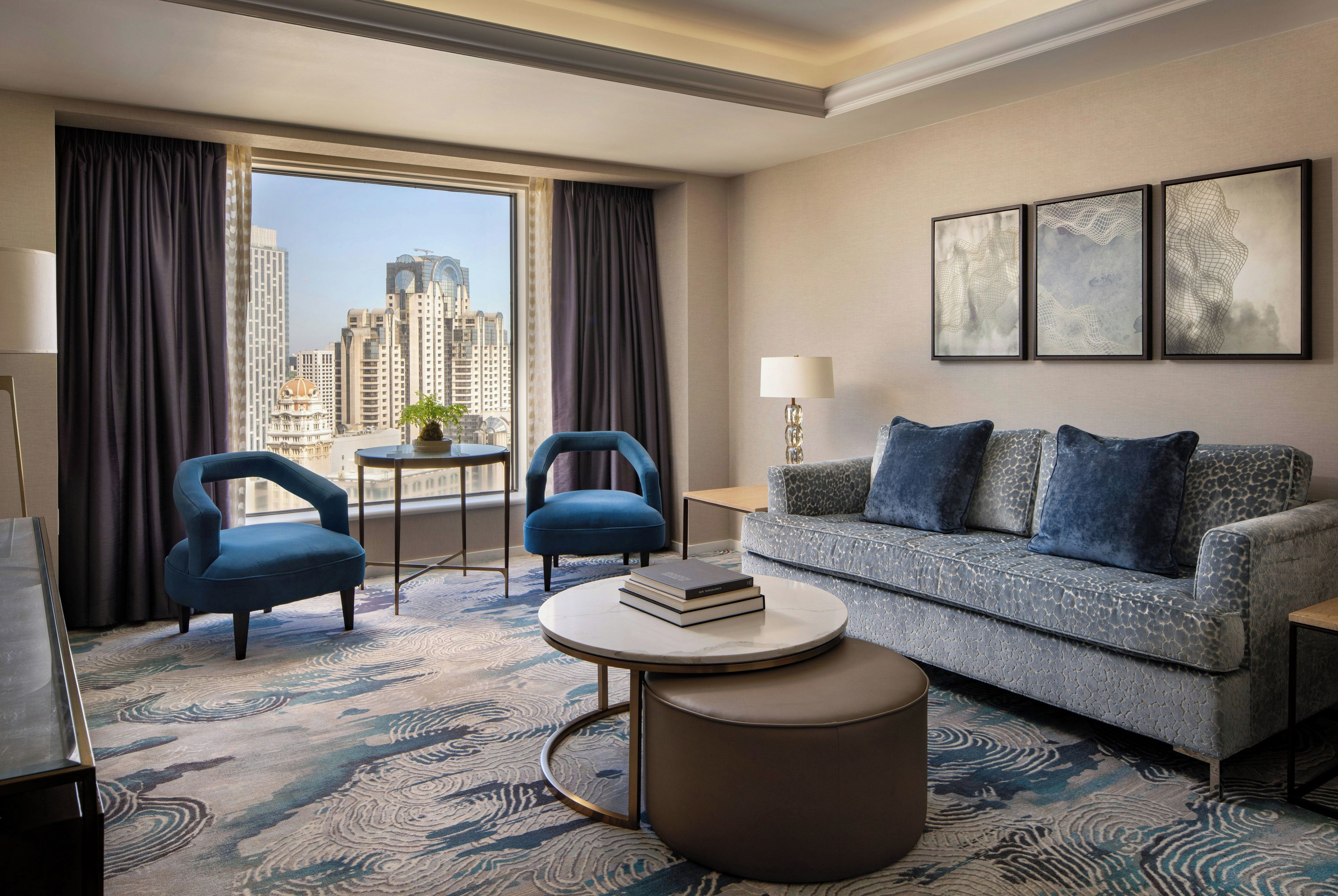 Ein Hotelzimmer mit Blick auf hohe Gebäude, zwei blaue Sessel, ein graues Sofa mit blauen Kissen, ein runder Couchtisch und eine Stehlampe.