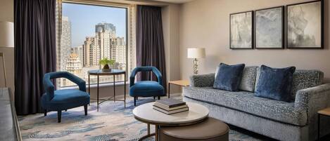 Ein Hotelzimmer mit Blick auf hohe Gebäude, zwei blaue Sessel, ein graues Sofa mit blauen Kissen, ein runder Couchtisch und eine Stehlampe.