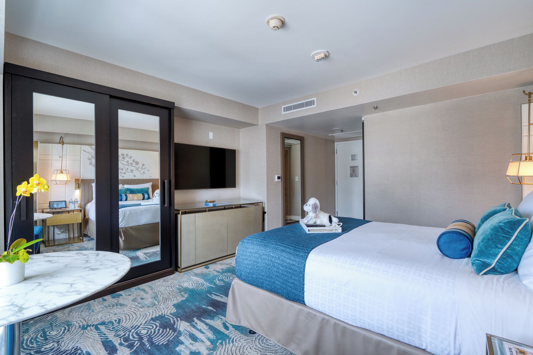 A hotel room with a large bed, a desk, a chair, a mirror, and a small table with a plant.