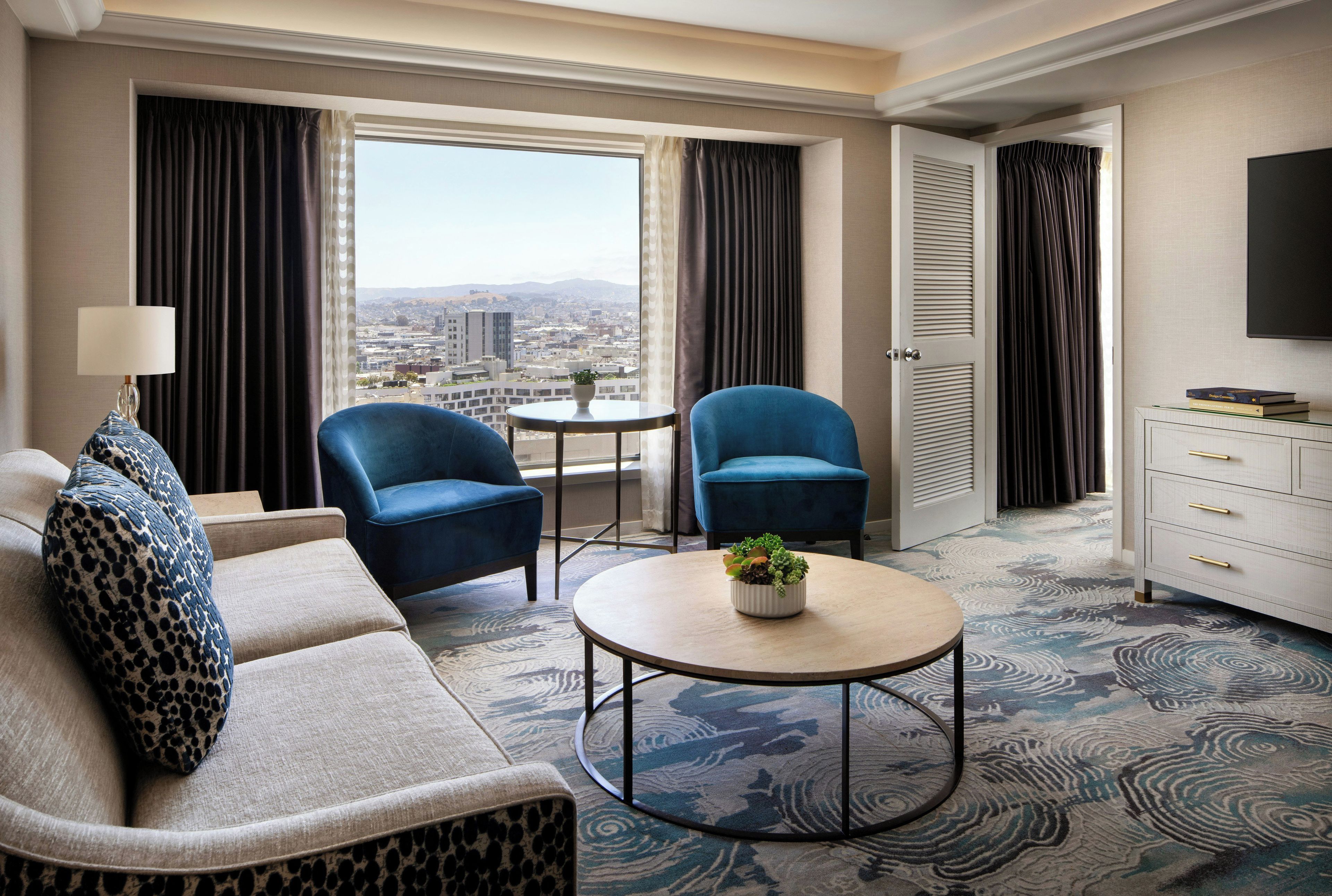 A hotel room with a sofa, two armchairs, a small table with a plant, a TV, and a view of the city.