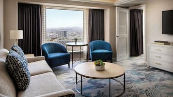 A hotel room with a sofa, two armchairs, a small table with a plant, a TV, and a view of the city.