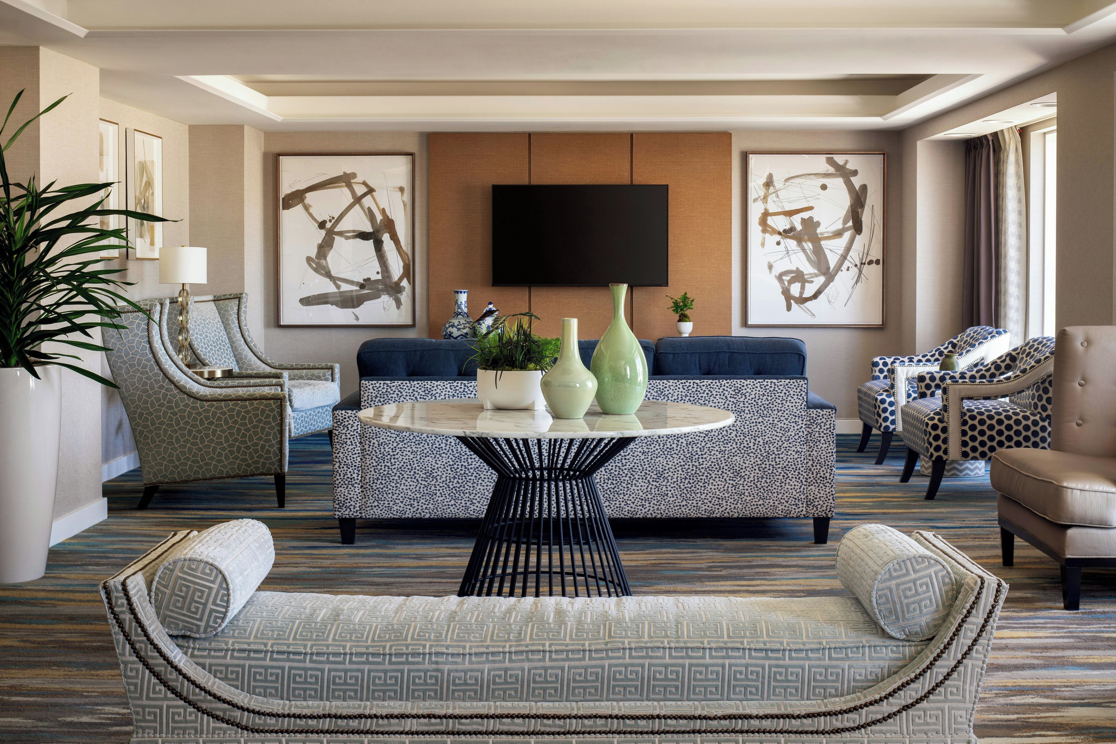 A modern living room with a round table, sofas, and chairs, a flat-screen TV mounted on the wall, and abstract art pieces.