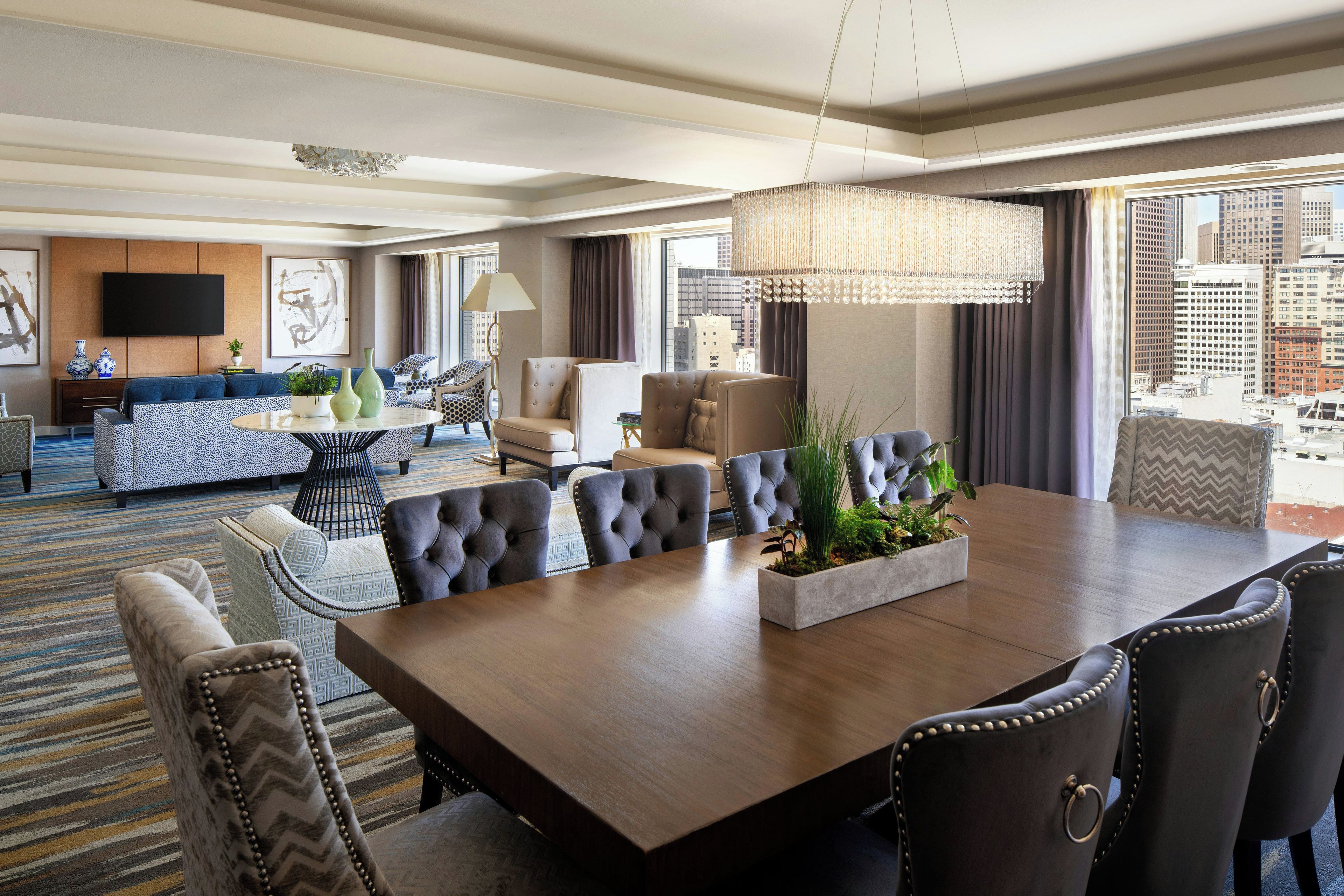 A hotel room with a dining area, a sofa set, a TV, and a city view.