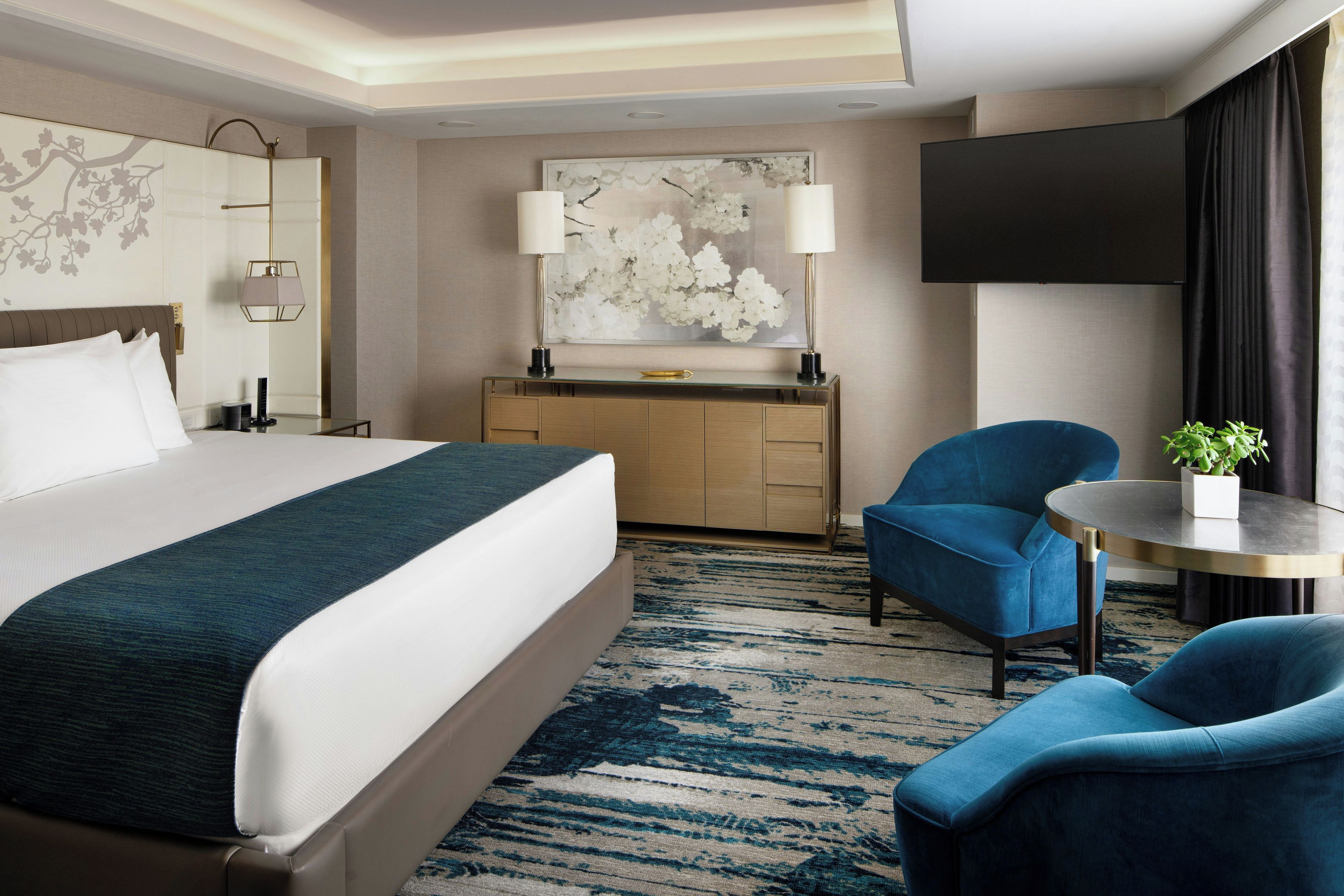 Habitación de hotel con cama, escritorio, dos sillas, una mesa redonda, televisor y un cuadro grande con motivos florales en la pared.