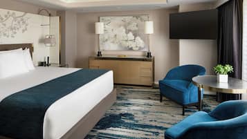 Habitación de hotel con cama, escritorio, dos sillas, una mesa redonda, televisor y un cuadro grande con motivos florales en la pared.