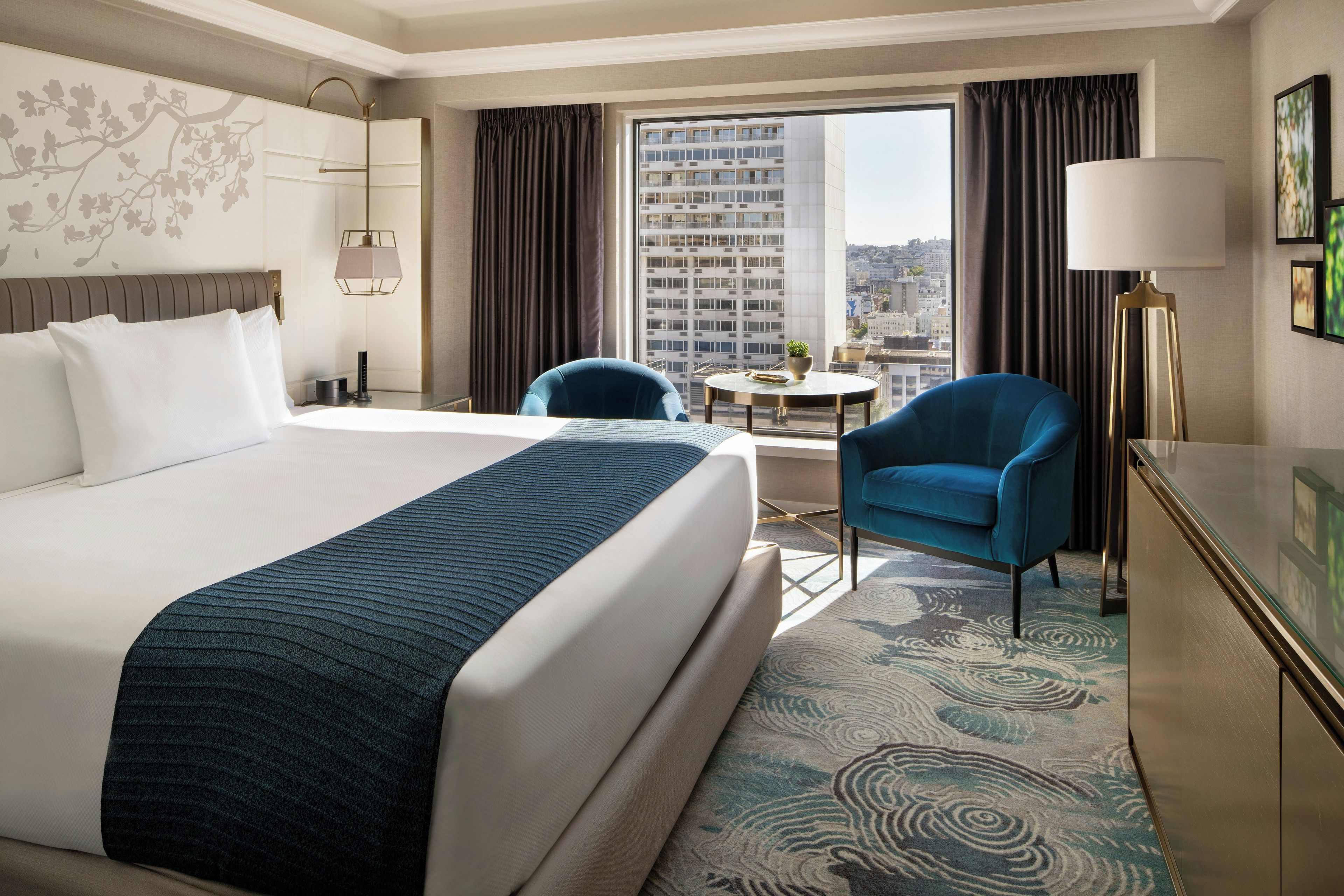 A hotel room with a large bed, a desk, a chair, a round table, and a view of a cityscape through the window.