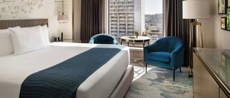 A hotel room with a large bed, a desk, a chair, a round table, and a view of a cityscape through the window.