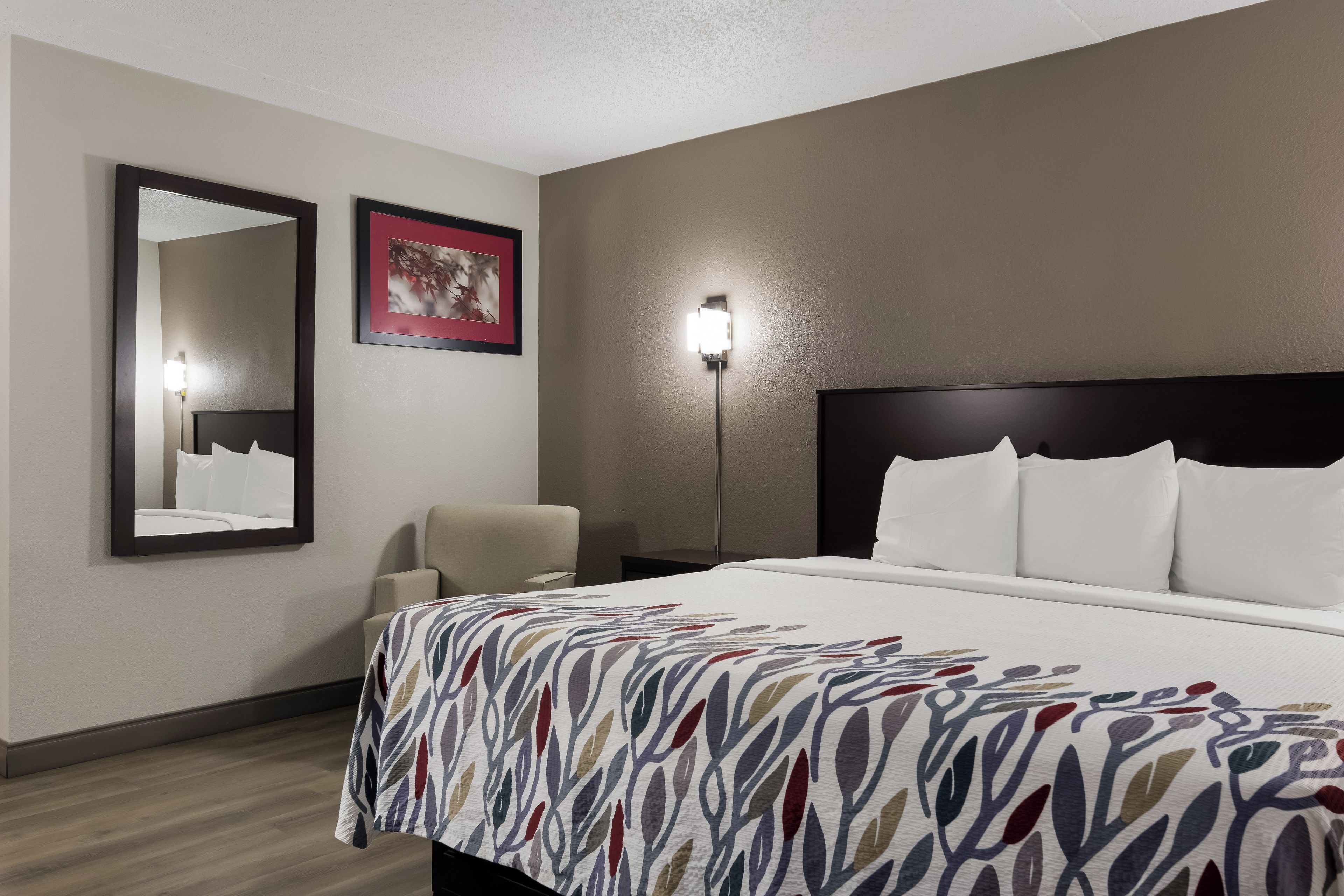 Ein Hotelzimmer mit einem großen Bett, einem Sessel, einem Nachttisch, einem wandmontierten Spiegel und einem gerahmten Bild.