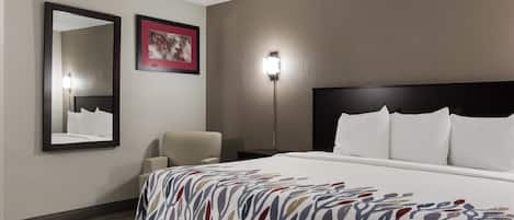 Une chambre d’hôtel avec un grand lit, une chaise, une table de chevet, un miroir fixé au mur et un tableau encadré.