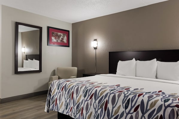 Una habitación de hotel con una cama grande, una silla, una mesita de noche, un espejo instalado en la pared y un cuadro enmarcado.