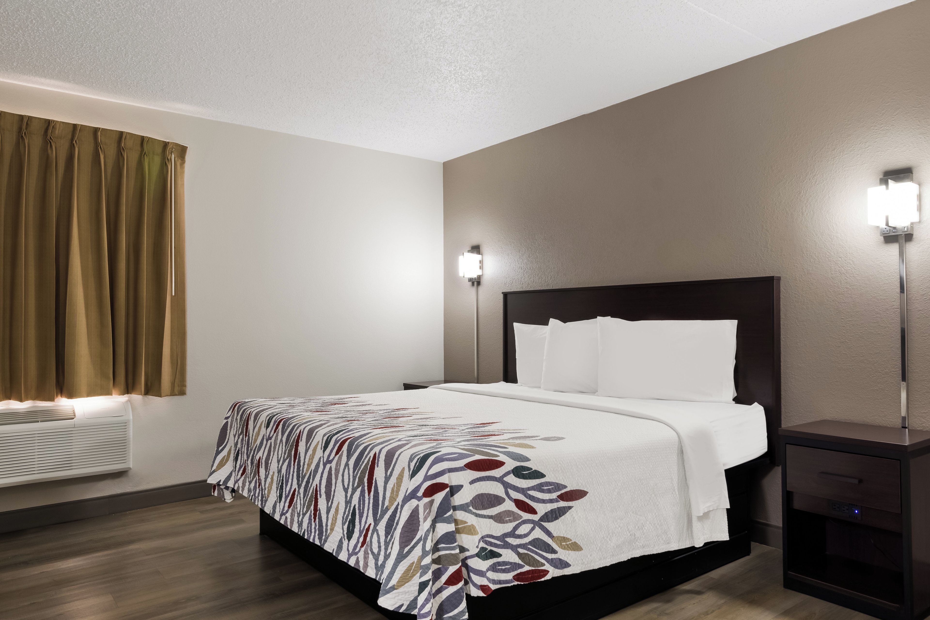 Quarto de hotel com cama, mesas de cabeceira, luminária na parede e janela com cortinas.