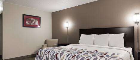 A hotel room with a bed, a chair, a nightstand, a lamp, and a framed picture on the wall.