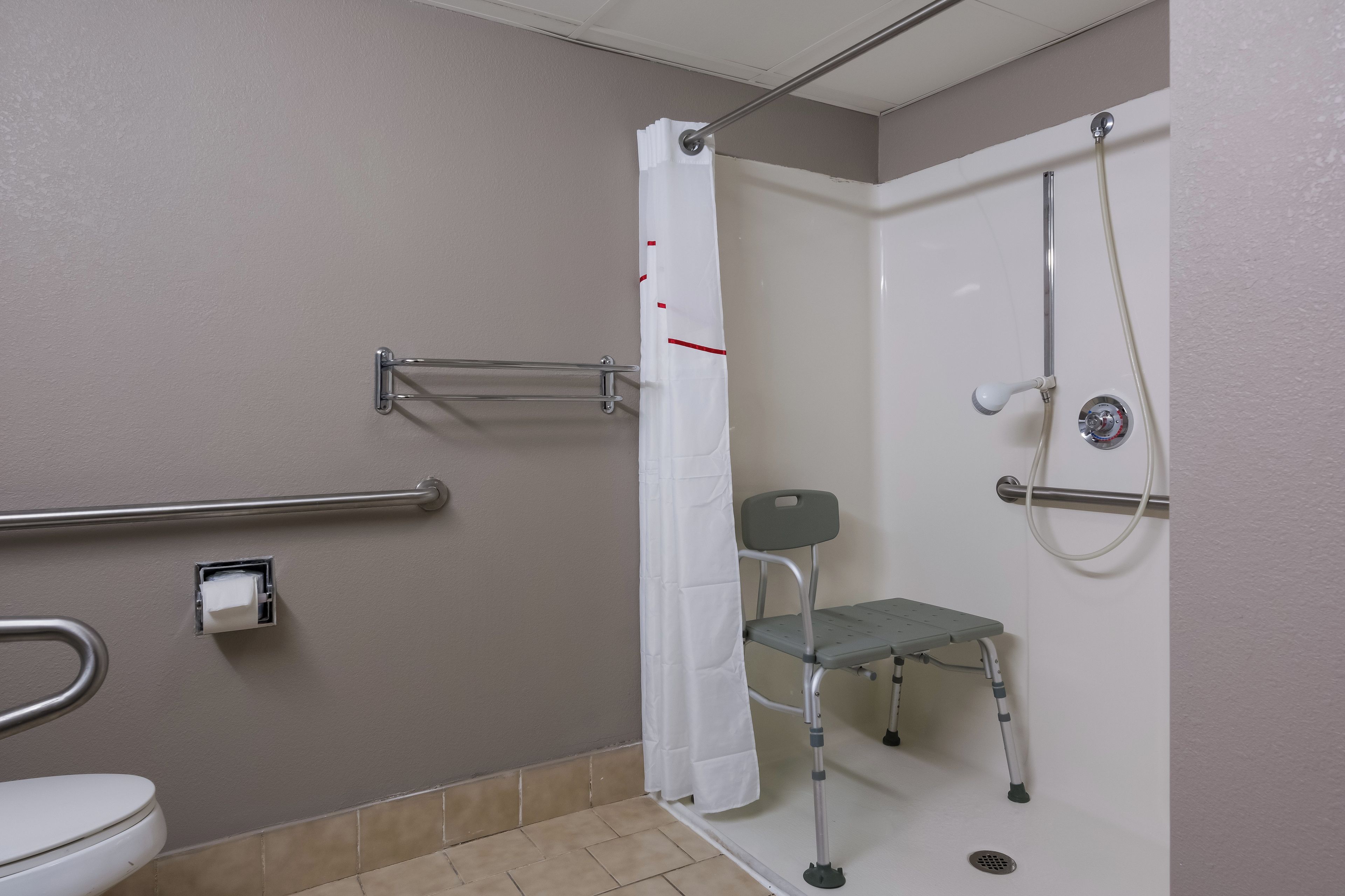 Un baño con inodoro, ducha con barra de apoyo, banco accesible para sillas de ruedas y secador de manos.