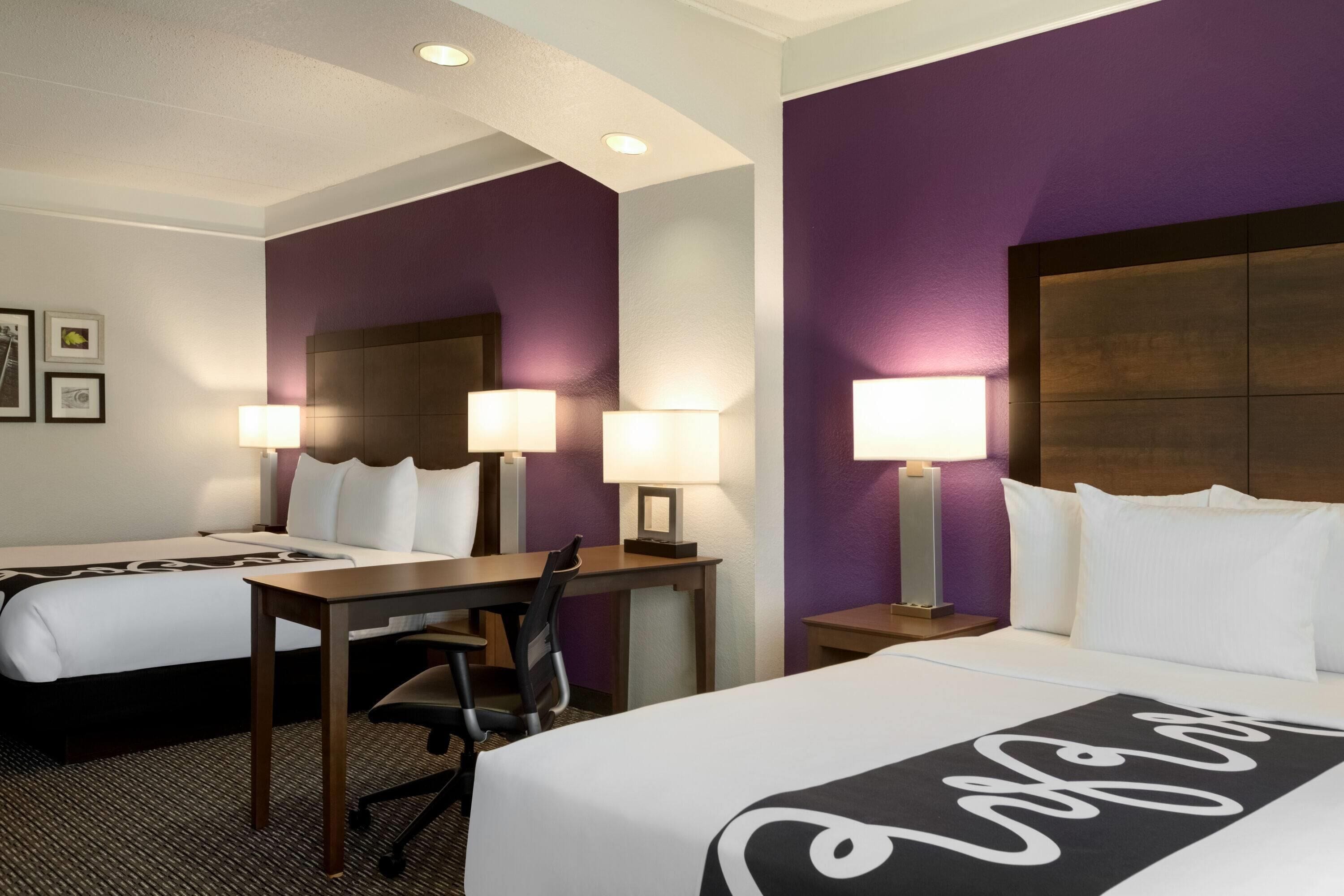 A hotel room with two beds, a desk, and a chair. There are wall-mounted lamps and a ceiling light.