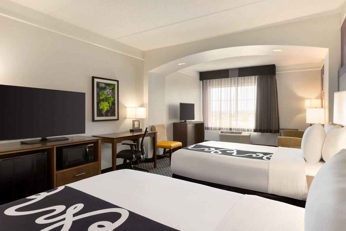 Habitación de hotel con dos camas, un escritorio con una silla, una televisión, un televisor de pantalla plana, una ventana con cortinas y un televisor montado en la pared.