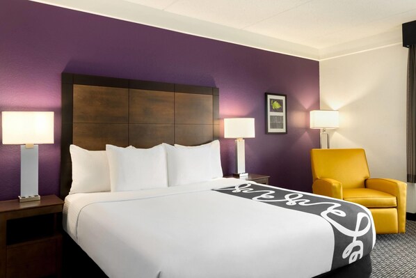 A hotel room with a large bed, two bedside lamps, a nightstand, a yellow armchair, and a purple wall.