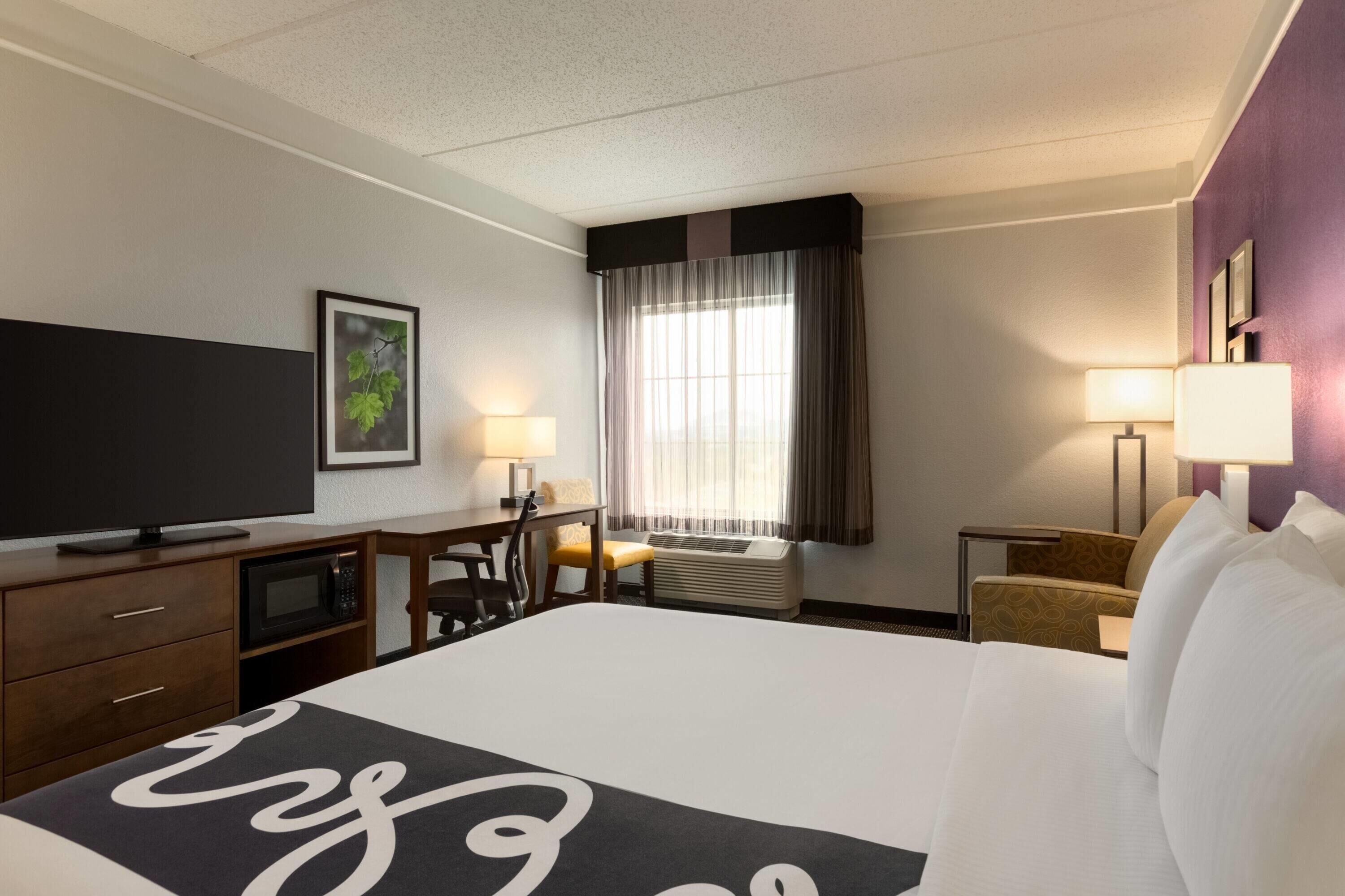 Una camera d'albergo con un letto, una scrivania, una televisione, una finestra con tende e una lampada a parete.