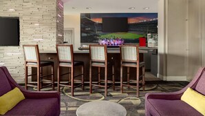 Habitación de hotel con comedor que presenta un gran mural de un estadio de béisbol, un televisor de pantalla plana montado en una pared de ladrillo y una zona de bar con una variedad de botellas.
