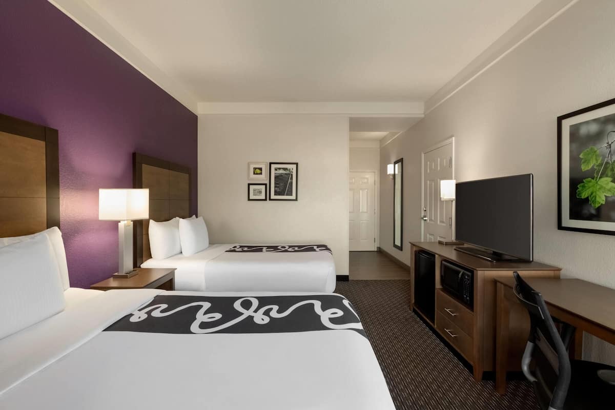 Habitación de hotel con dos camas, un escritorio, un televisor y una puerta que da a un pasillo.