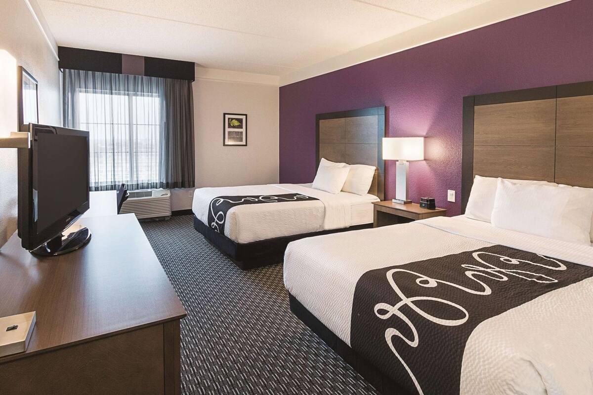 Habitación de hotel con dos camas, un escritorio con televisor, una ventana con cortinas y una lámpara fijada a la pared.