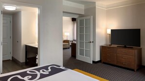 Habitación de hotel con una cama, un televisor sobre un aparador de madera, un escritorio con una silla y una puerta que da a otra habitación.