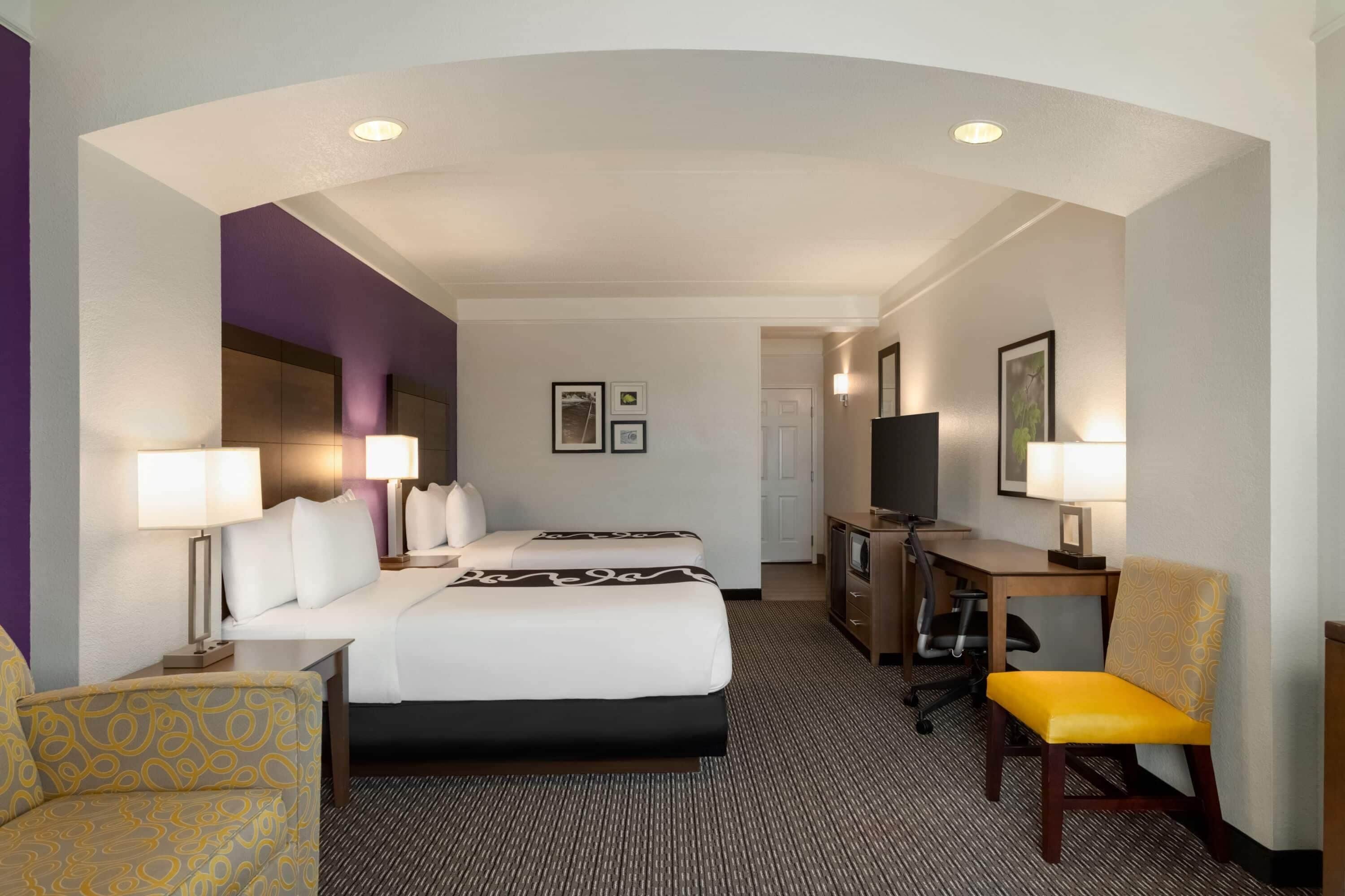 A hotel room with a bed, two bedside tables, a desk with a chair, a TV, and a patterned armchair.