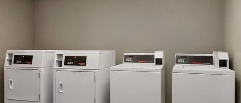 A row of four white commercial washers in a room with beige walls and a tiled ceiling.