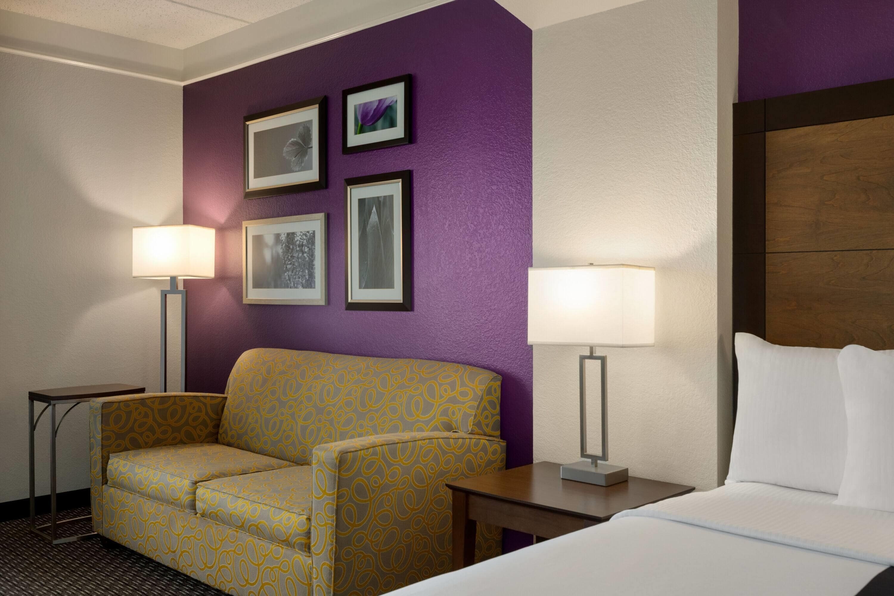 A hotel room with a bed, a sofa, two lamps, and framed pictures on the wall.